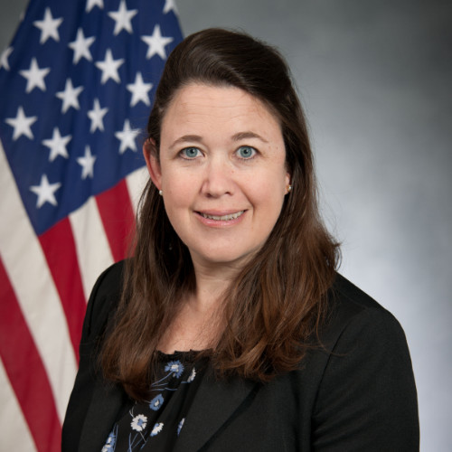 image of Heather wearing a black suit standing in front of an American flag, smiling at the camera