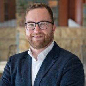 headshot of Lukas wearing glasses and a blue blazer smiling at the camera