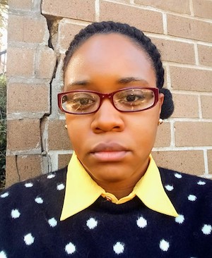 headshot of Menika weaing glasses standing against a brick wall in a blue and white polka dot sweater and yellow collar shirt