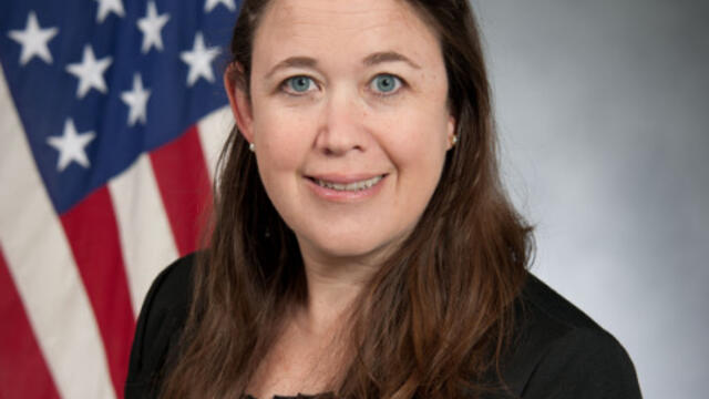 image of Heather wearing a black suit standing in front of an American flag, smiling at the camera