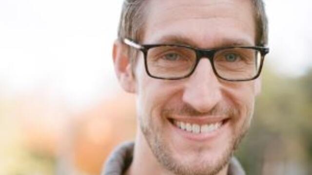 image of Jacob standing outside wearing glasses and a tan sweater smiling at the camera