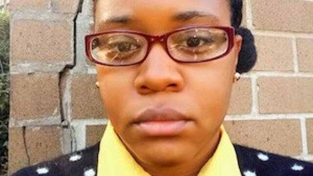 headshot of Menika weaing glasses standing against a brick wall in a blue and white polka dot sweater and yellow collar shirt