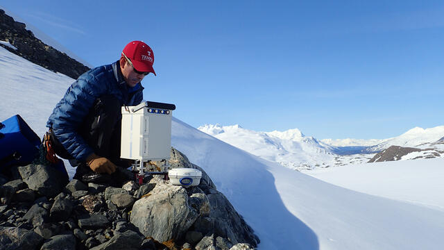 A man in a red Temple baseball cap perched on rocks with a snowy backdrop.