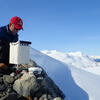 A man in a red Temple baseball cap perched on rocks with a snowy backdrop.