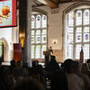 Image of a speaker standing at a podium in a cathedral-like hall, addressing an audience.. A large screen to the left shows a close-up of hands holding an apple and a decorated donu