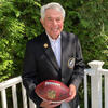 Merrill Reese holding a football and wearing his Eagles Hall of Fame jacket outside.