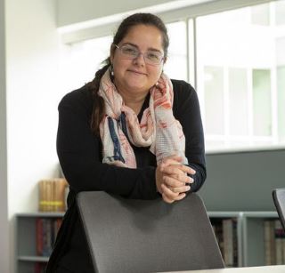 image of Muge smiling and leaning on a chair wearing glasses a black sweater and tan and peach scarf